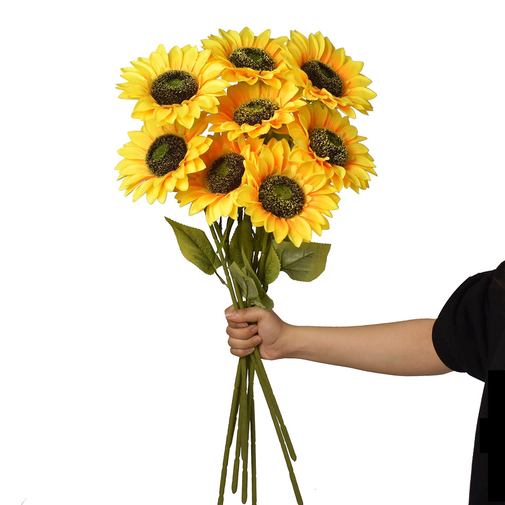 Hand holding a bouquet of 8 artificial sunflowers with green stems and leaves against a white background.