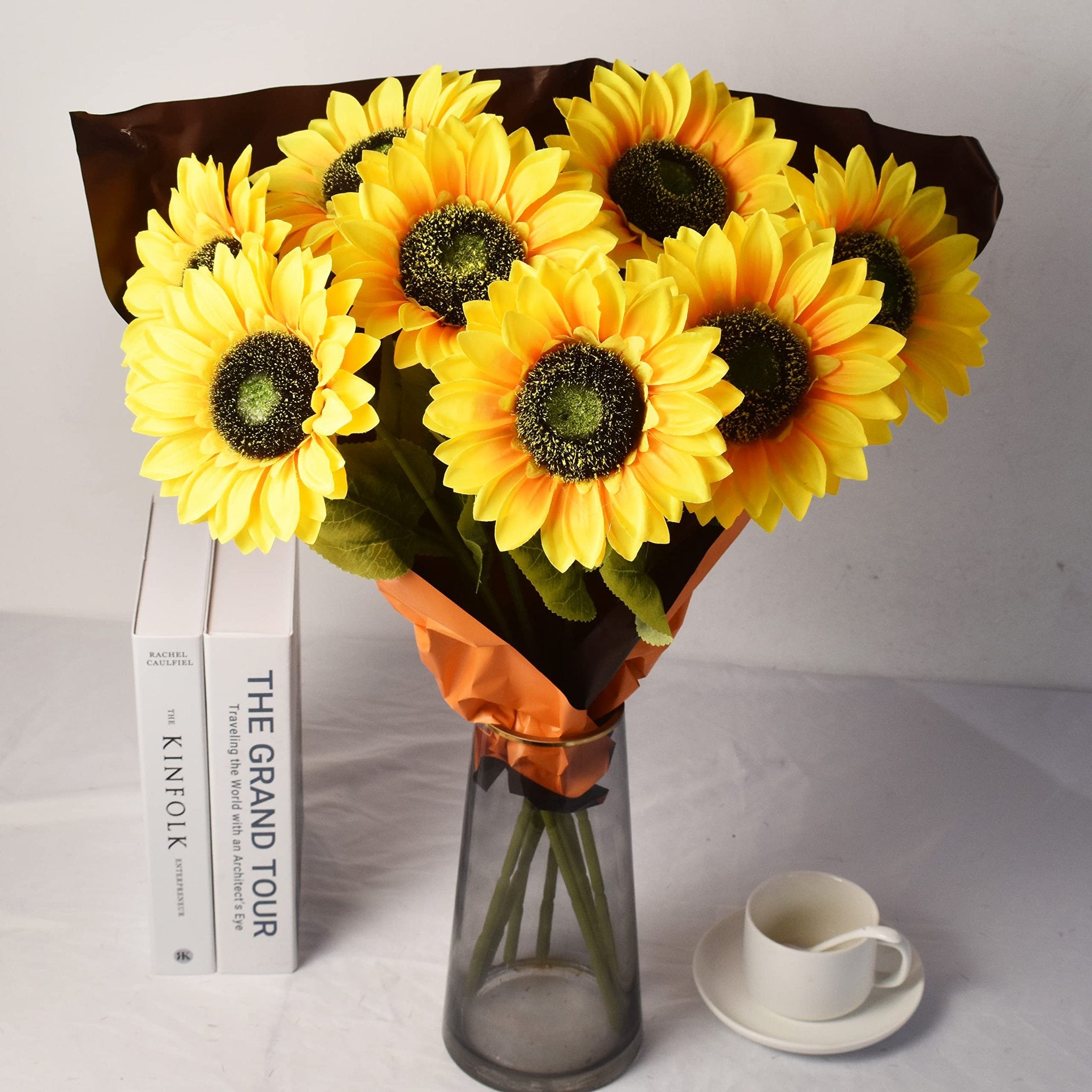 Bouquet of eight artificial sunflowers in a vase, with books and a teacup nearby.