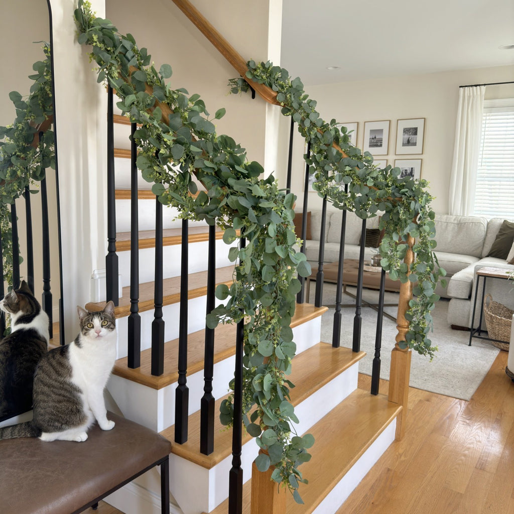 Artificial eucalyptus garland draped on a wooden staircase with black spindles, two cats sit on a bench nearby.