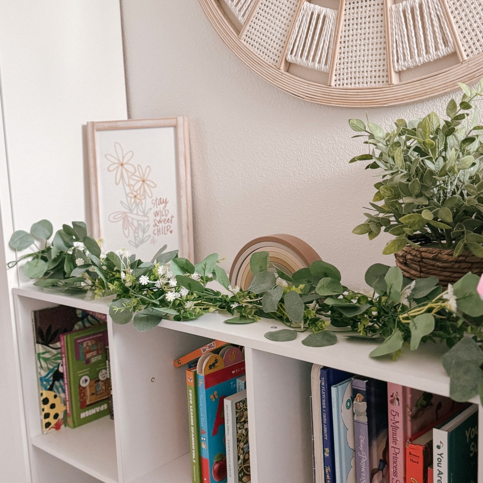 Artificial eucalyptus garland with silk leaves and small white flowers draped across a white bookshelf with books and decor.