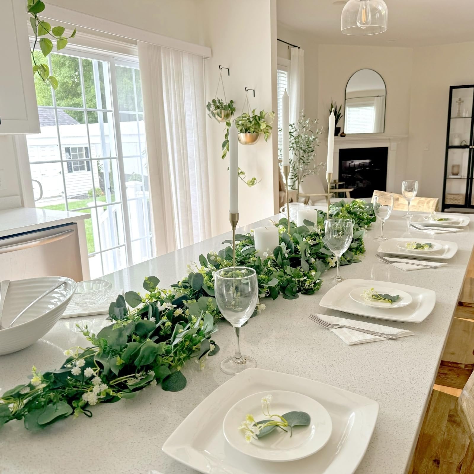Artificial eucalyptus garland with white flowers laid across a set dining table with place settings and candles.