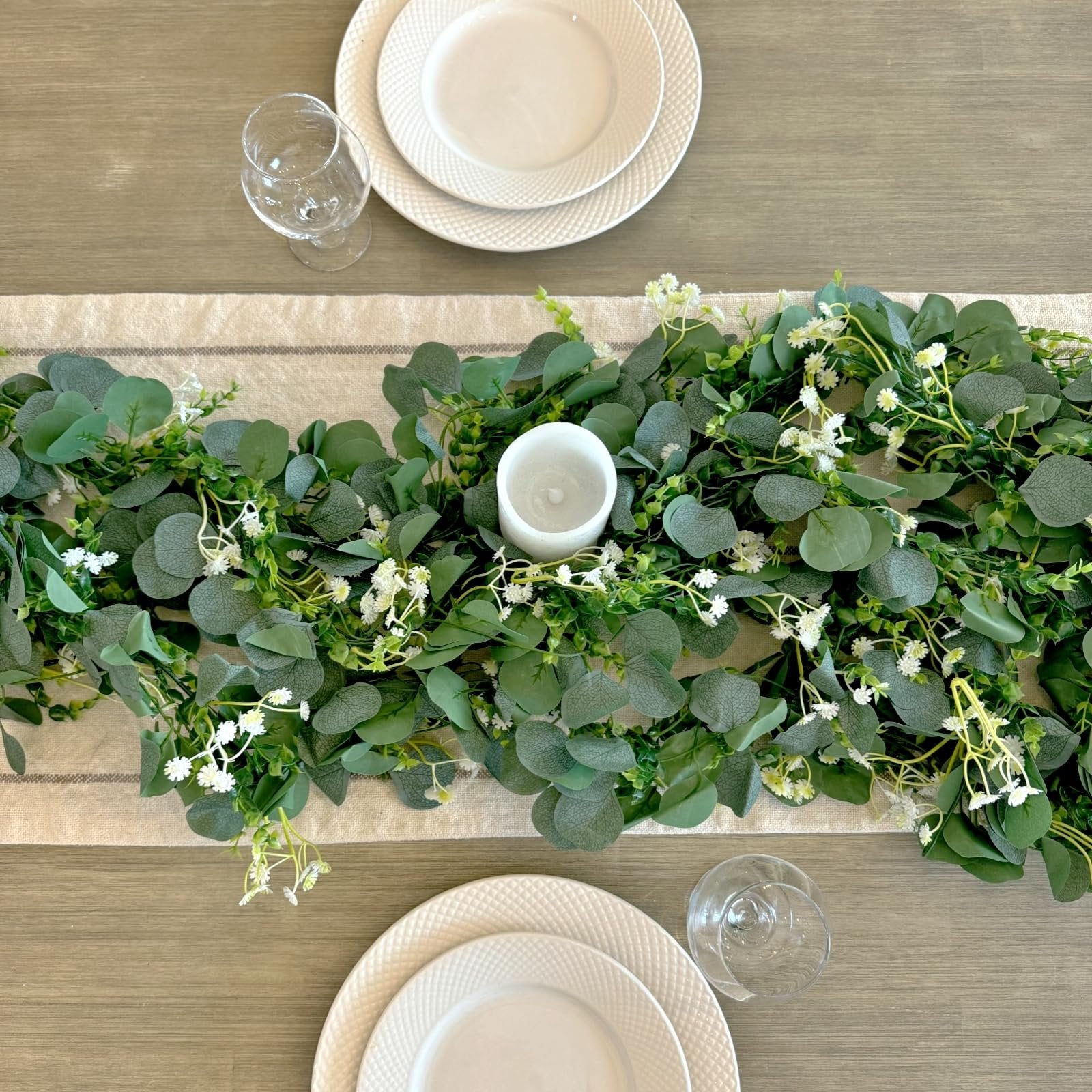 Artificial eucalyptus garland with white flowers draped across a table runner with place settings and a candle
