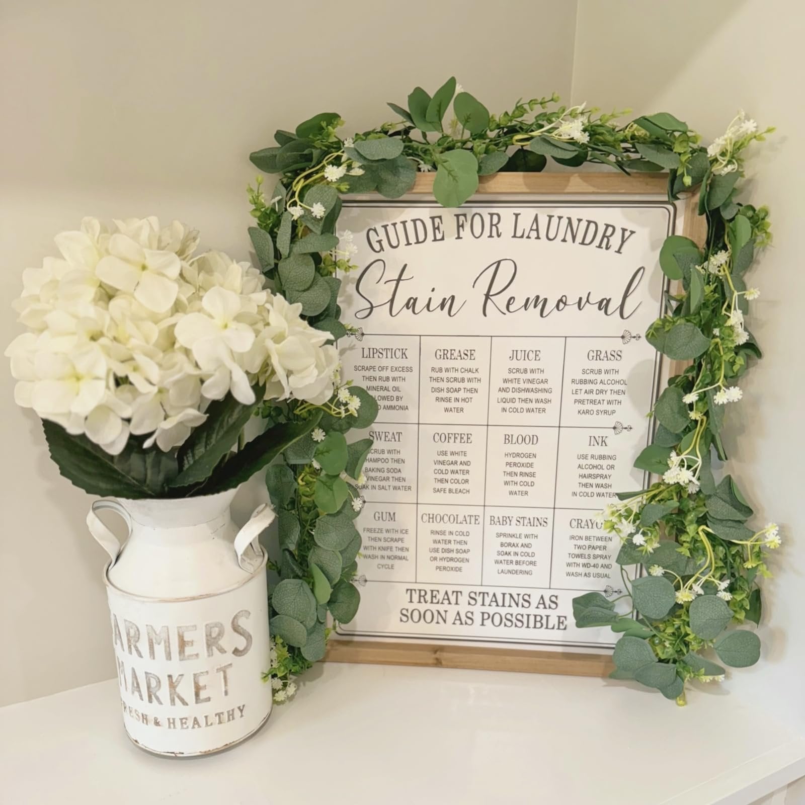 Guide for laundry stain removal sign surrounded by artificial eucalyptus garland and white hydrangeas in a distressed white jug.