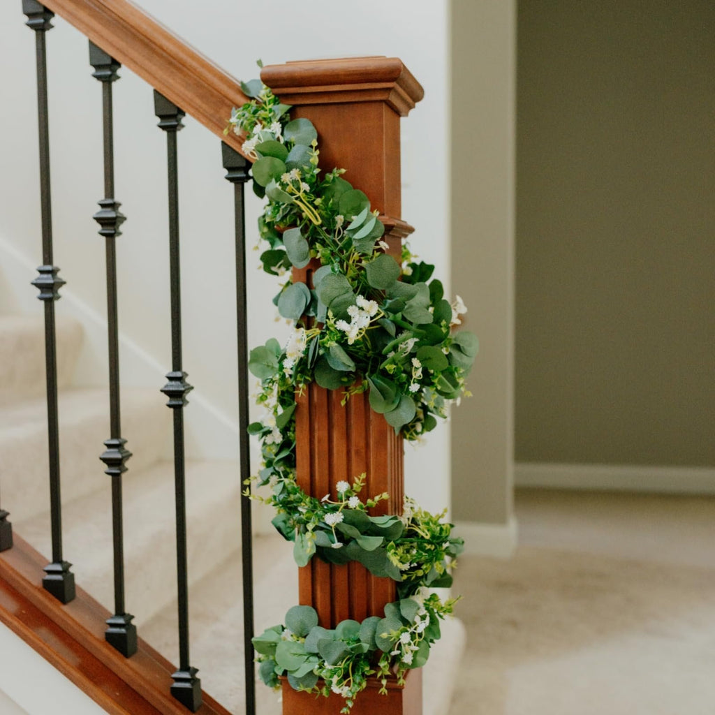 Artificial eucalyptus garland with silk leaves and small white flowers draped around a wooden newel post on a staircase.