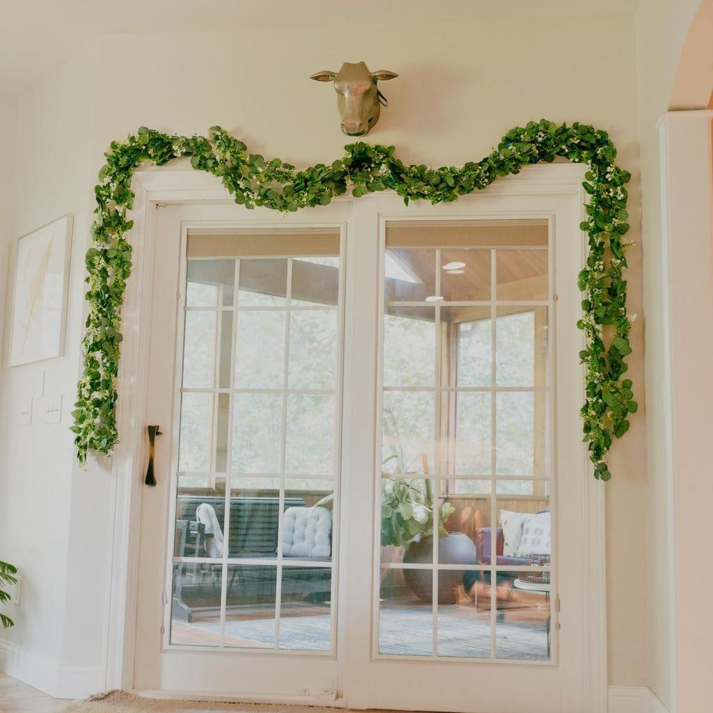 Artificial eucalyptus garland with silk leaves draped over a doorway, featuring a brass cow head mounted above.