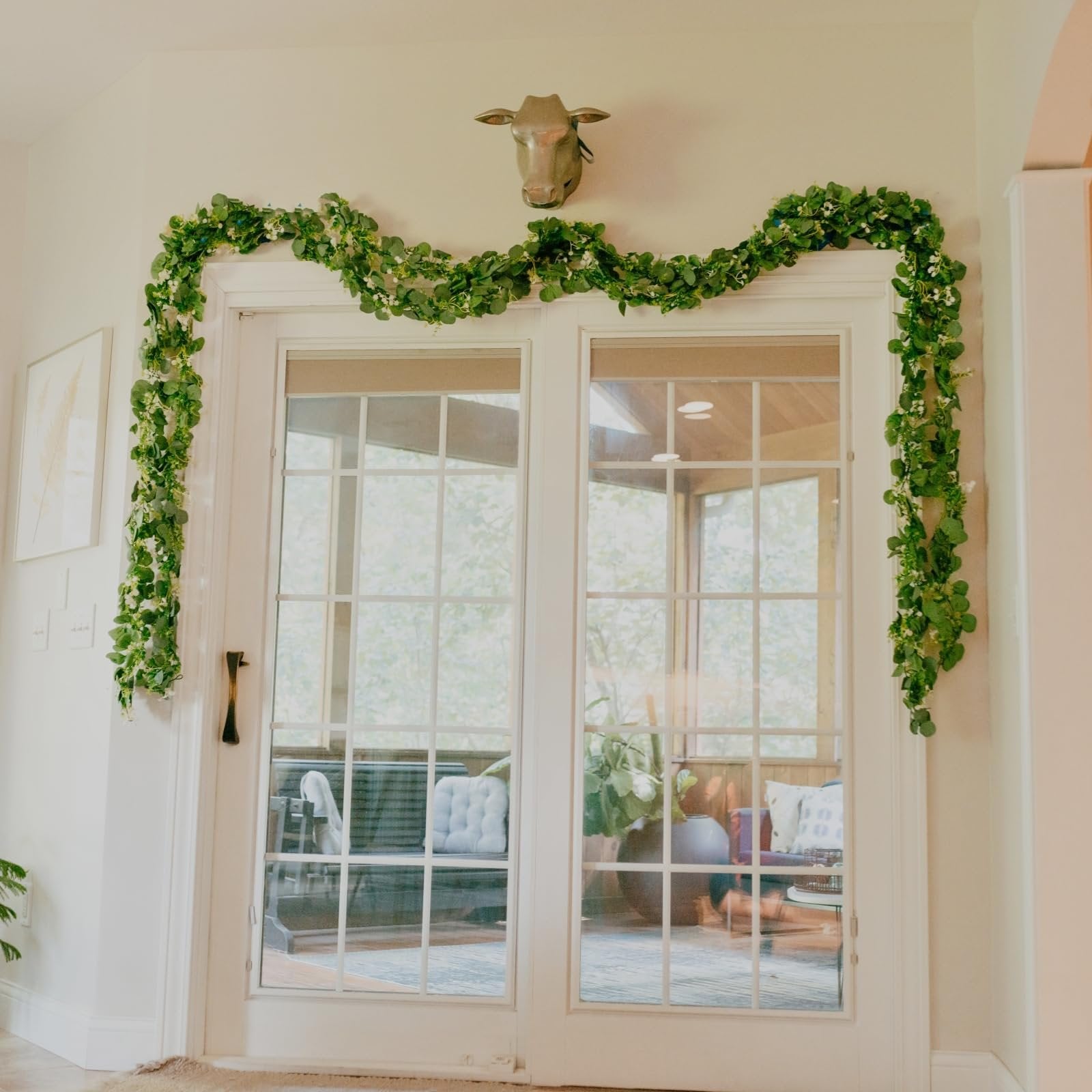 Artificial eucalyptus garland with silk leaves draped over a doorway, featuring a brass cow head mounted above.