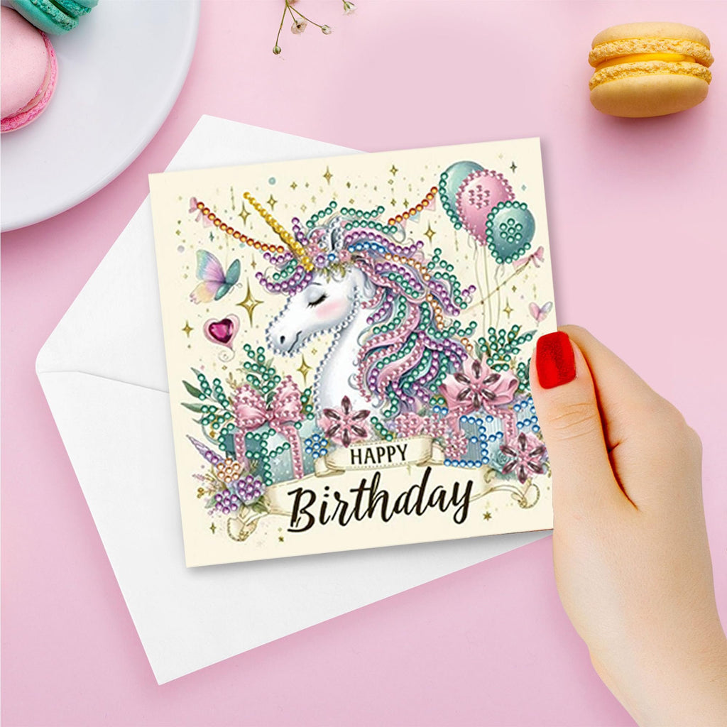 Hand holding a diamond painting birthday card featuring a unicorn, balloons, and gifts, with macarons and a plate in the background.