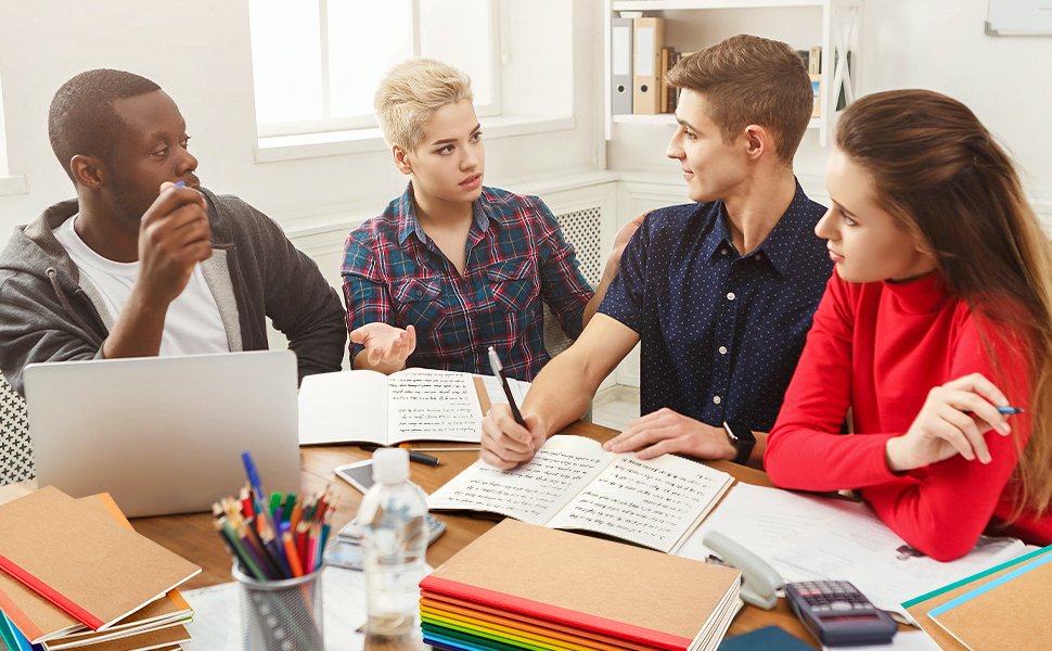 Four young adults collaborating around a table with notebooks and a laptop, discussing studies.
