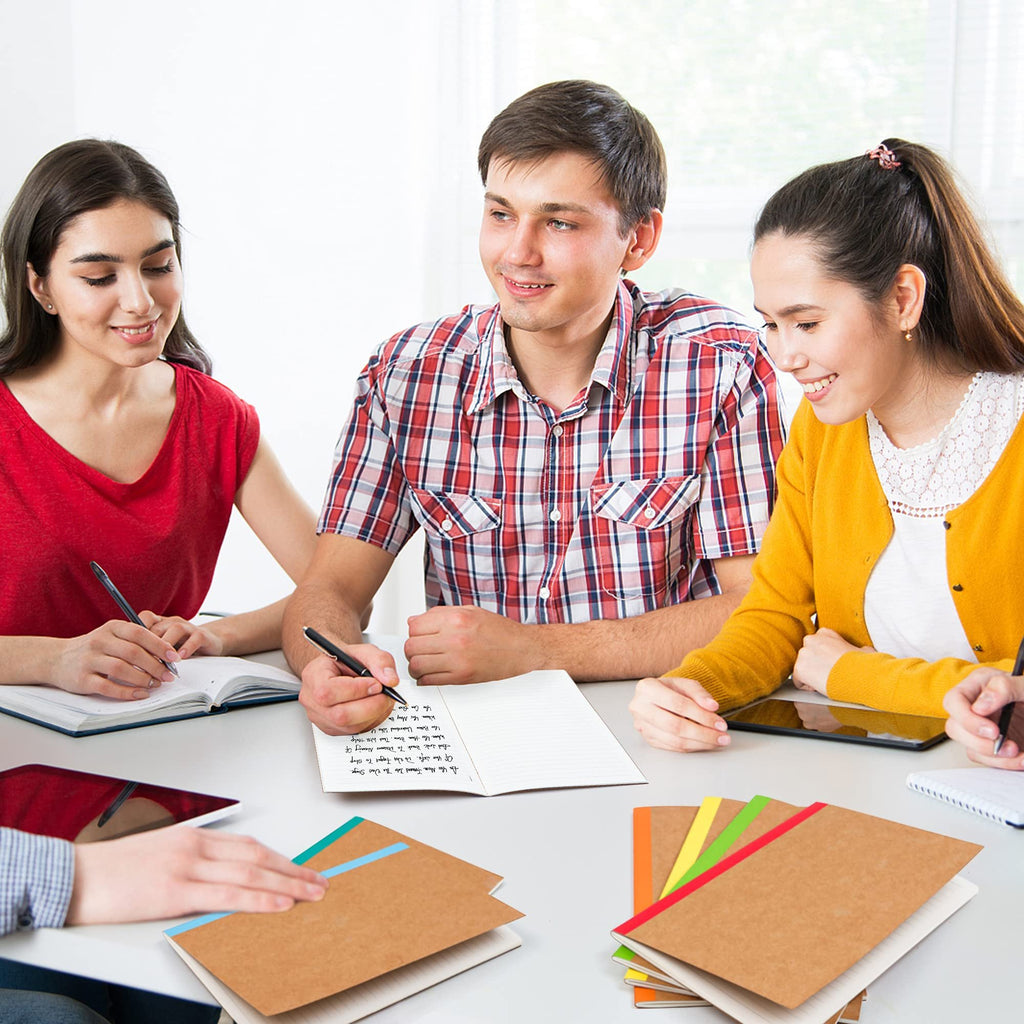 Four students studying at a table with notebooks and tablets, one student writes in a notebook.