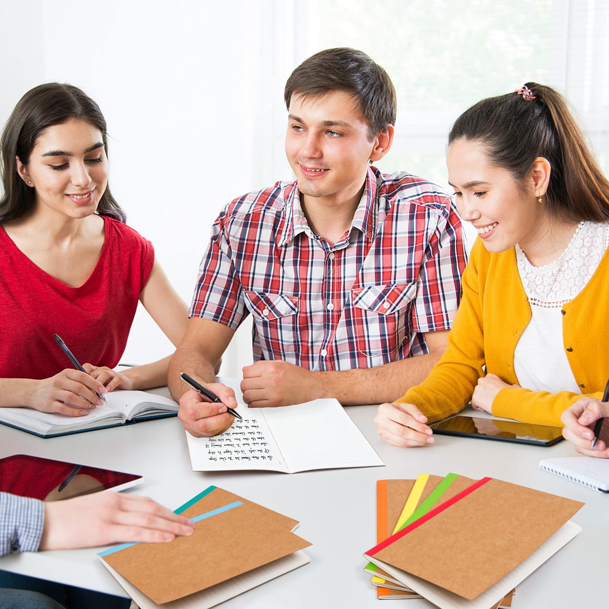 Four students studying at a table with notebooks and tablets, one student writes in a notebook.