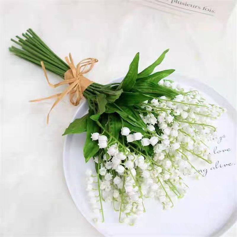 Bouquet of artificial lily of the valley flowers with green leaves tied with twine on a white plate.
