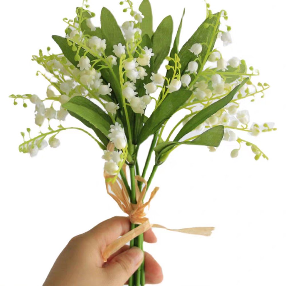Hand holding a bouquet of artificial white Lily of the Valley flowers with green leaves, tied with a straw ribbon.