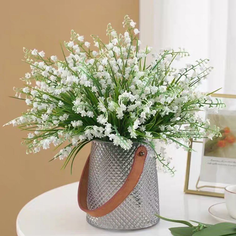 Bouquet of artificial white lily of the valley flowers in a textured gray vase with a leather handle.