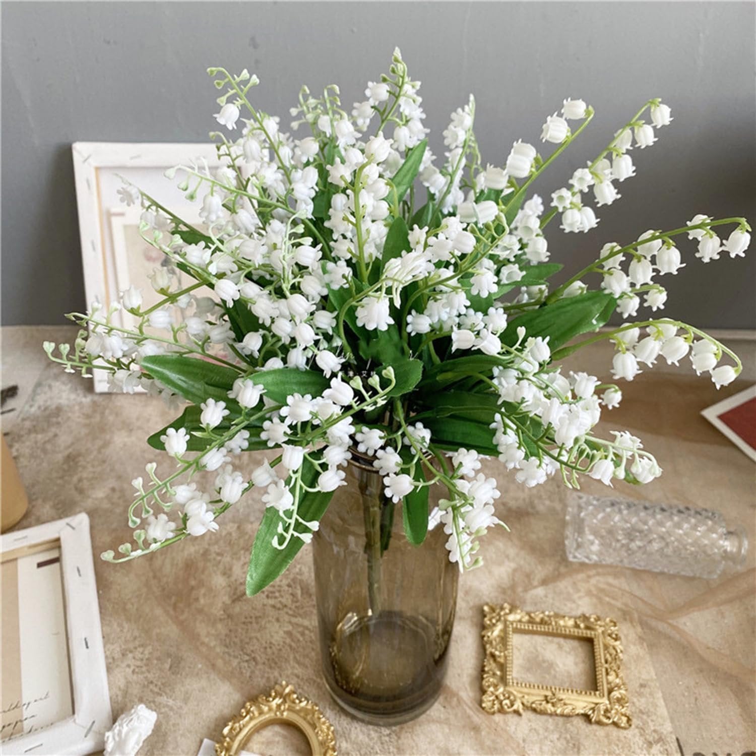 Bouquet of artificial white lily of the valley flowers in a brown glass vase, with decorative frames on the side.