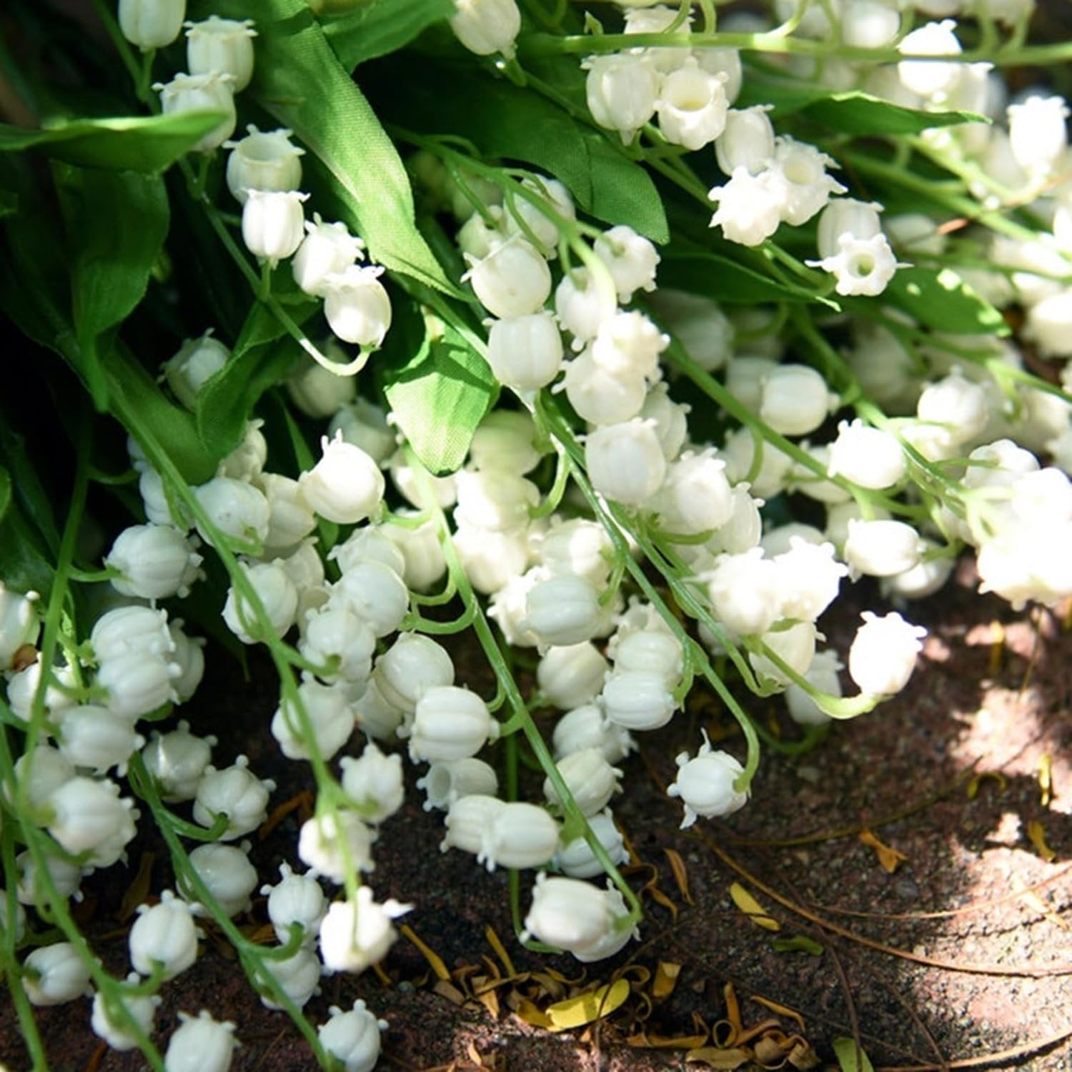 Close-up of a bunch of artificial white Lily of the Valley flowers with green stems and leaves, resting on a textured surface.