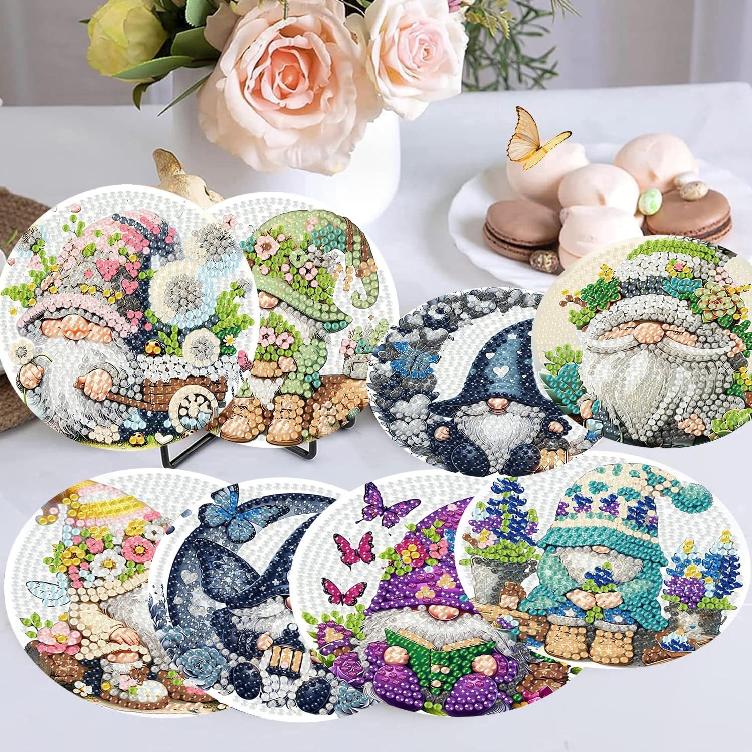 Eight diamond art coasters featuring gnomes and floral designs, arranged on a table with flowers and macarons in the background.