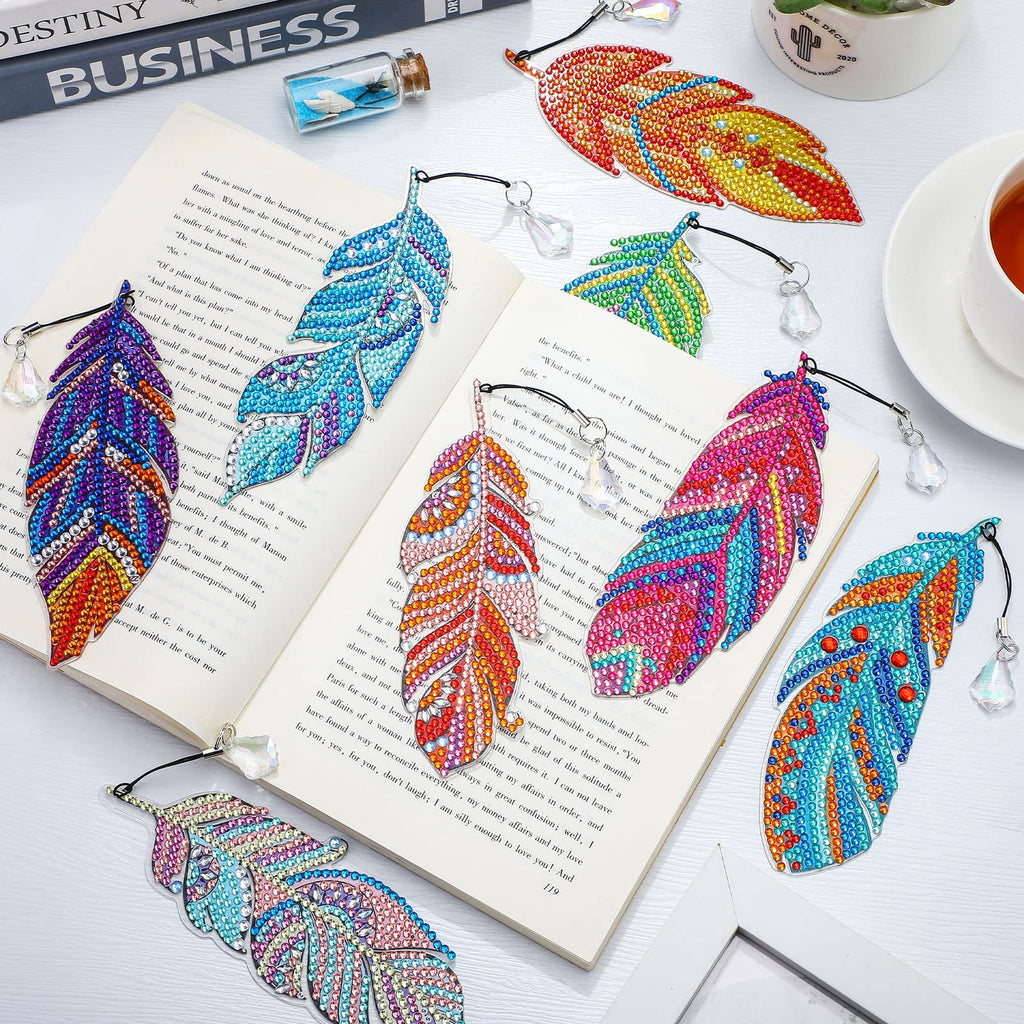 Eight colorful diamond art feather bookmarks arranged on an open book, with a cup of tea and decorative items in the background.