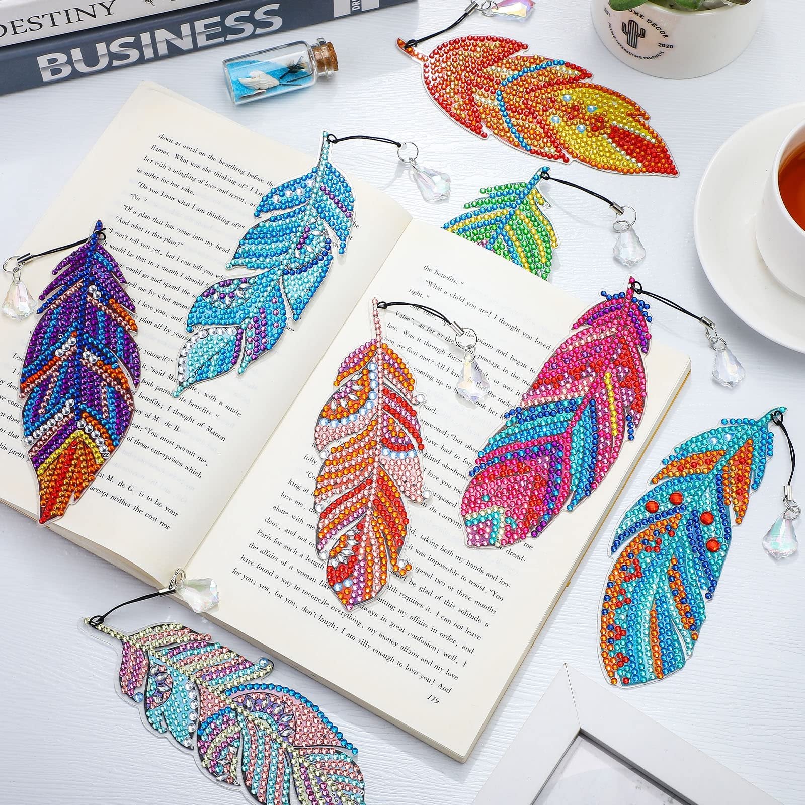 Eight colorful diamond art feather bookmarks arranged on an open book, with a cup of tea and decorative items in the background.