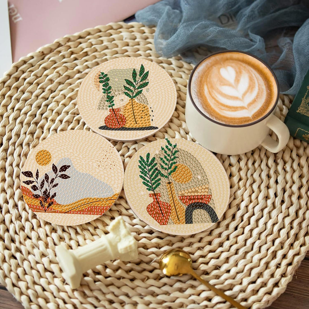 Three bohemian style diamond art coasters with abstract landscape and plant designs, arranged on a woven mat next to a cup of coffee.
