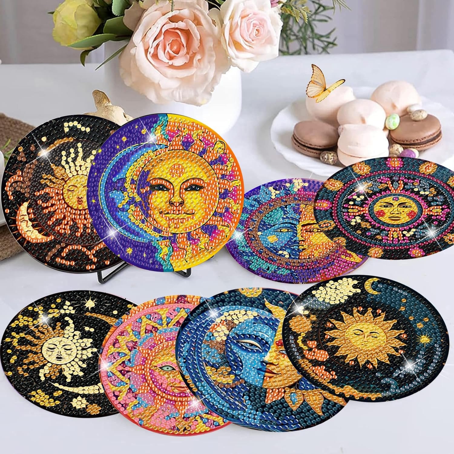 Eight diamond art coasters with sun and moon designs displayed on a table with flowers and pastries in the background.