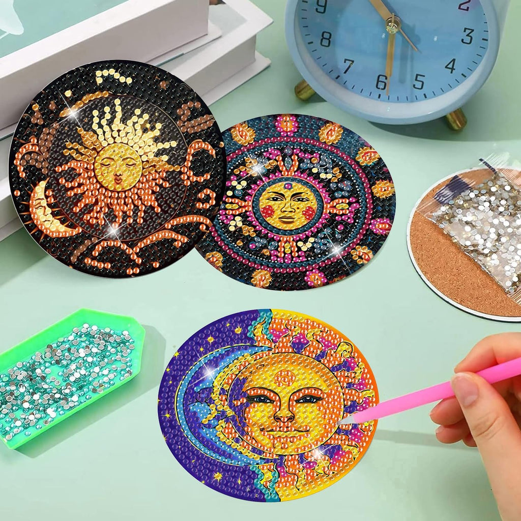 Diamond art coasters with sun and moon designs, one being worked on with a pink tool, next to a tray of gems and a bag of gems.