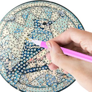 Hand holding pink diamond painting tool applying gems to a round coaster with a blue jay design