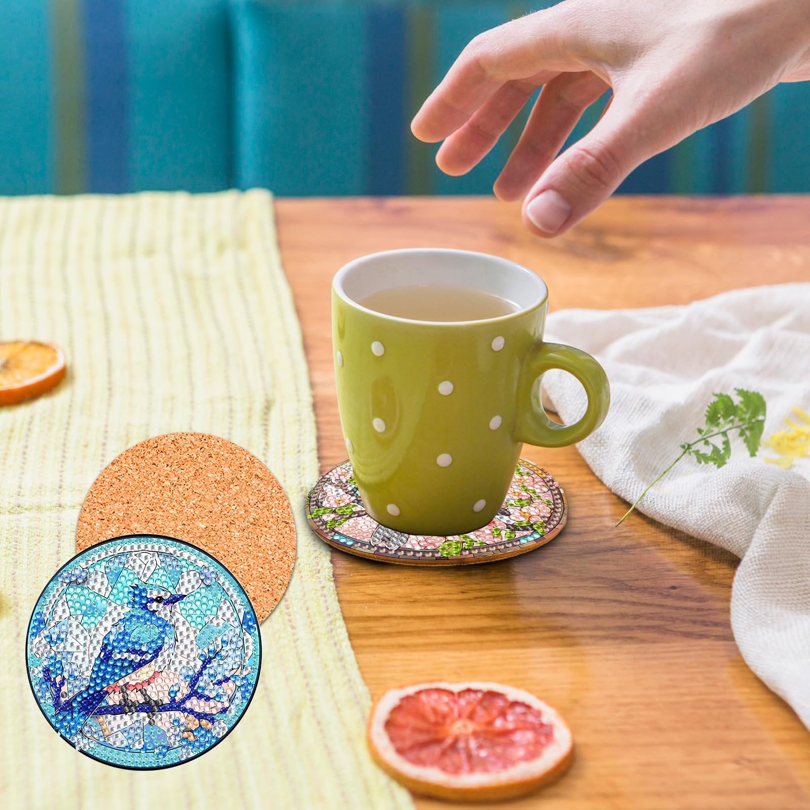 Diamond painting coasters with a blue jay and floral designs, one under a green mug on a wooden table.
