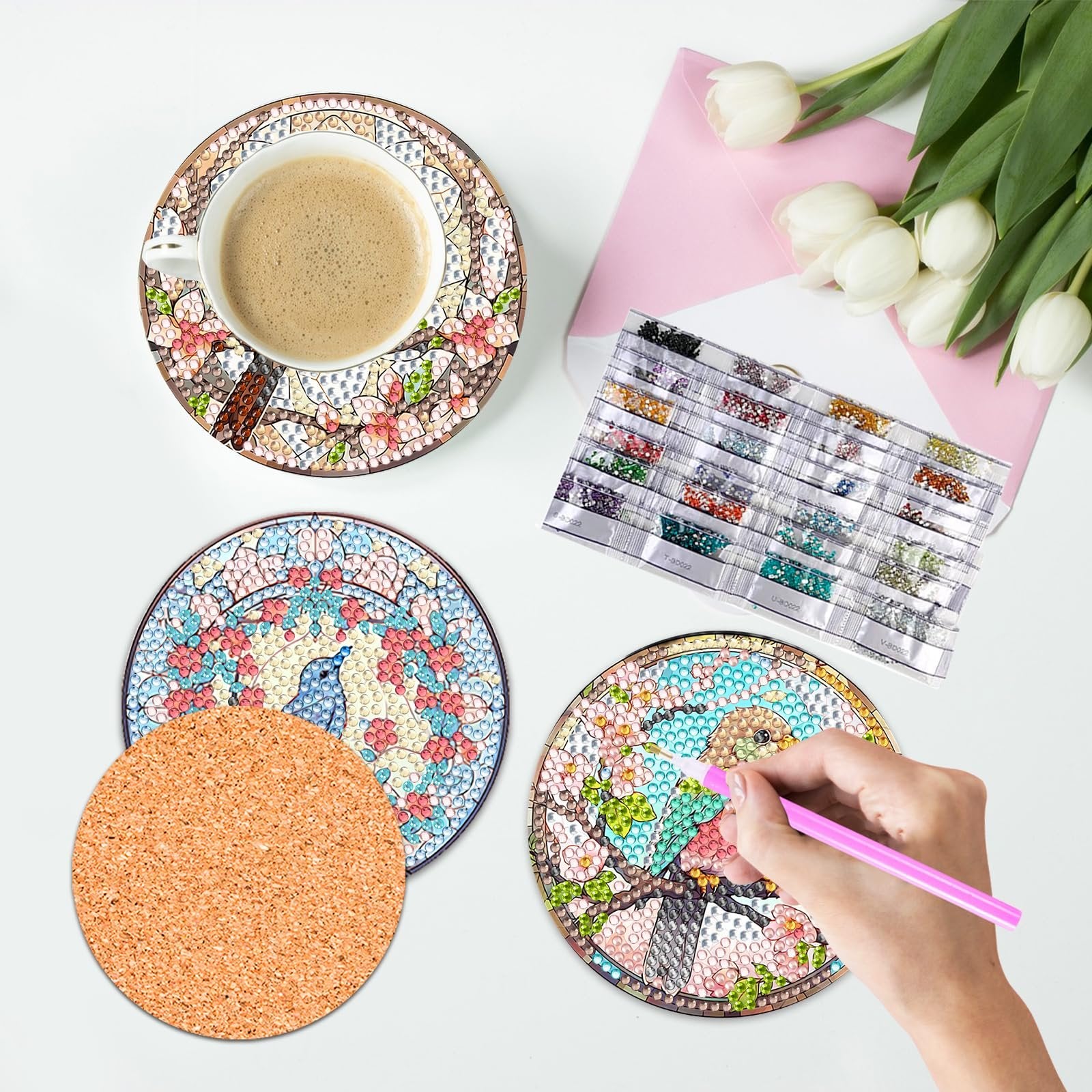 Diamond painting coasters kit with coffee cup, cork base, and diamond beads laid out on a white surface with tulips and pink envelope.