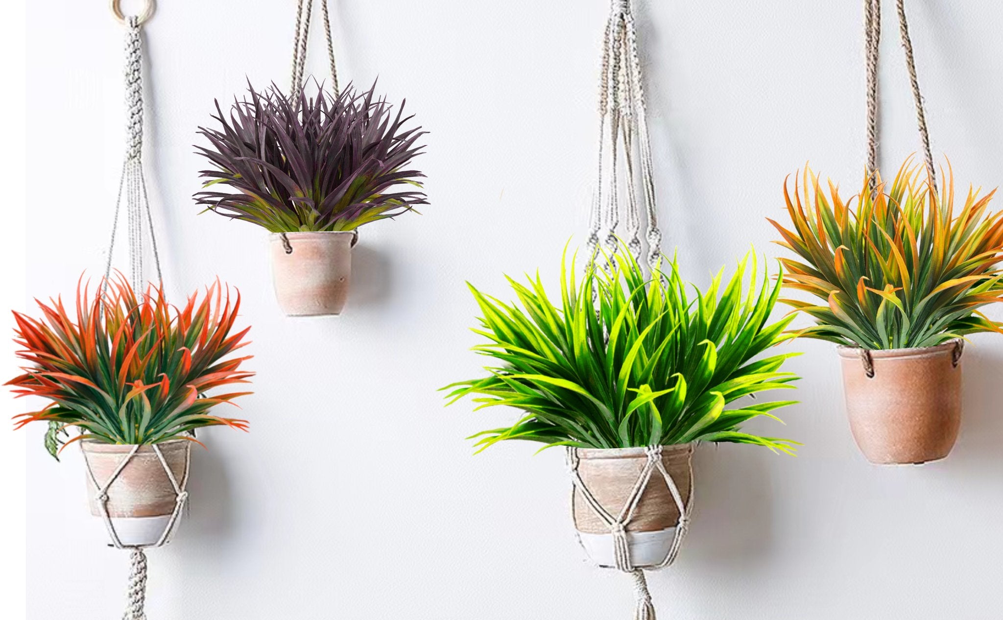 Four hanging artificial plants with macrame hangers against a white wall. Plants are in terracotta pots and feature green, orange, and purple foliage.