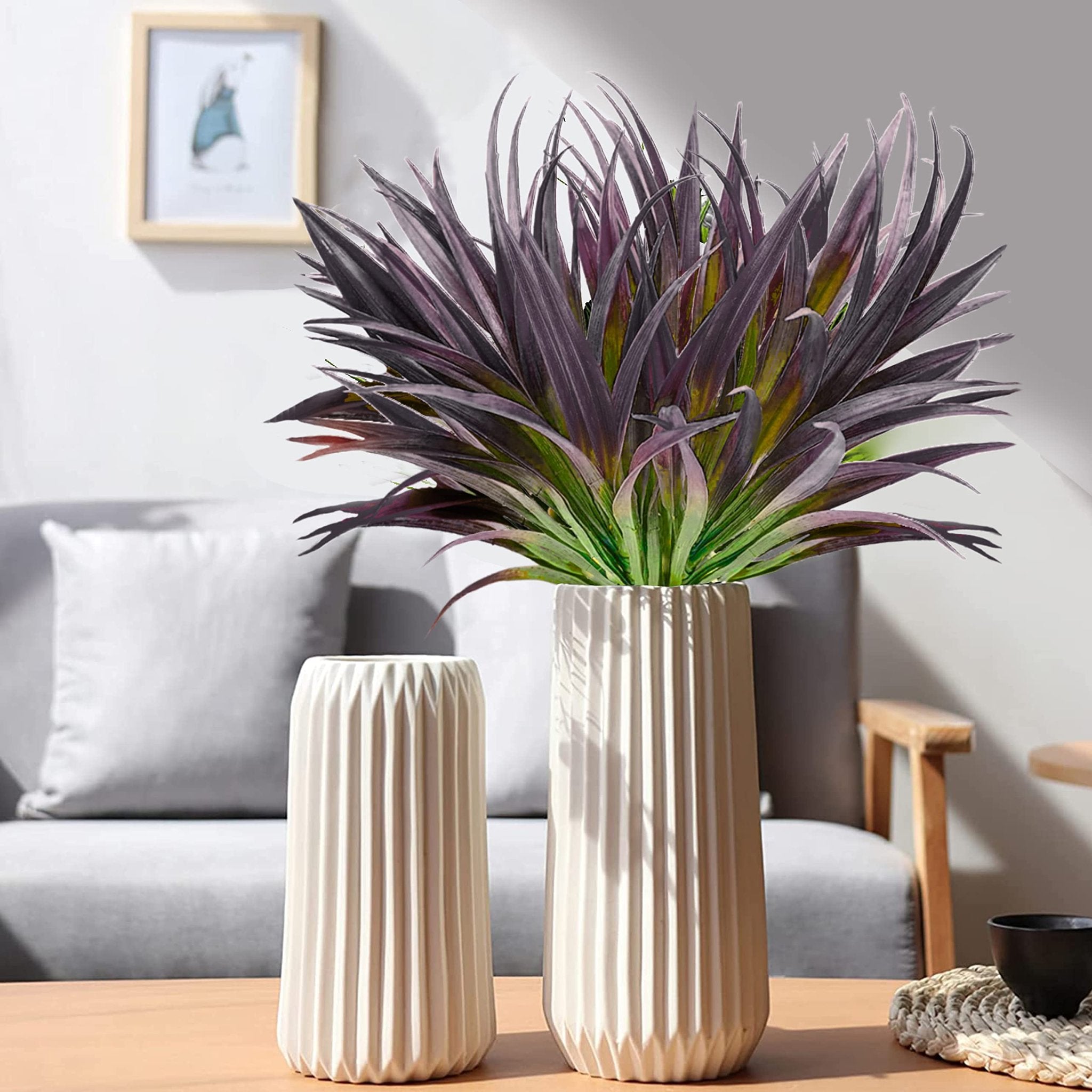 Two ribbed ceramic vases with artificial purple and green grass plants on a wooden table in a living room setting.