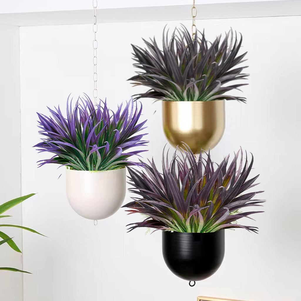Three hanging planters with artificial tall grass plants in purple and green hues against a white wall.