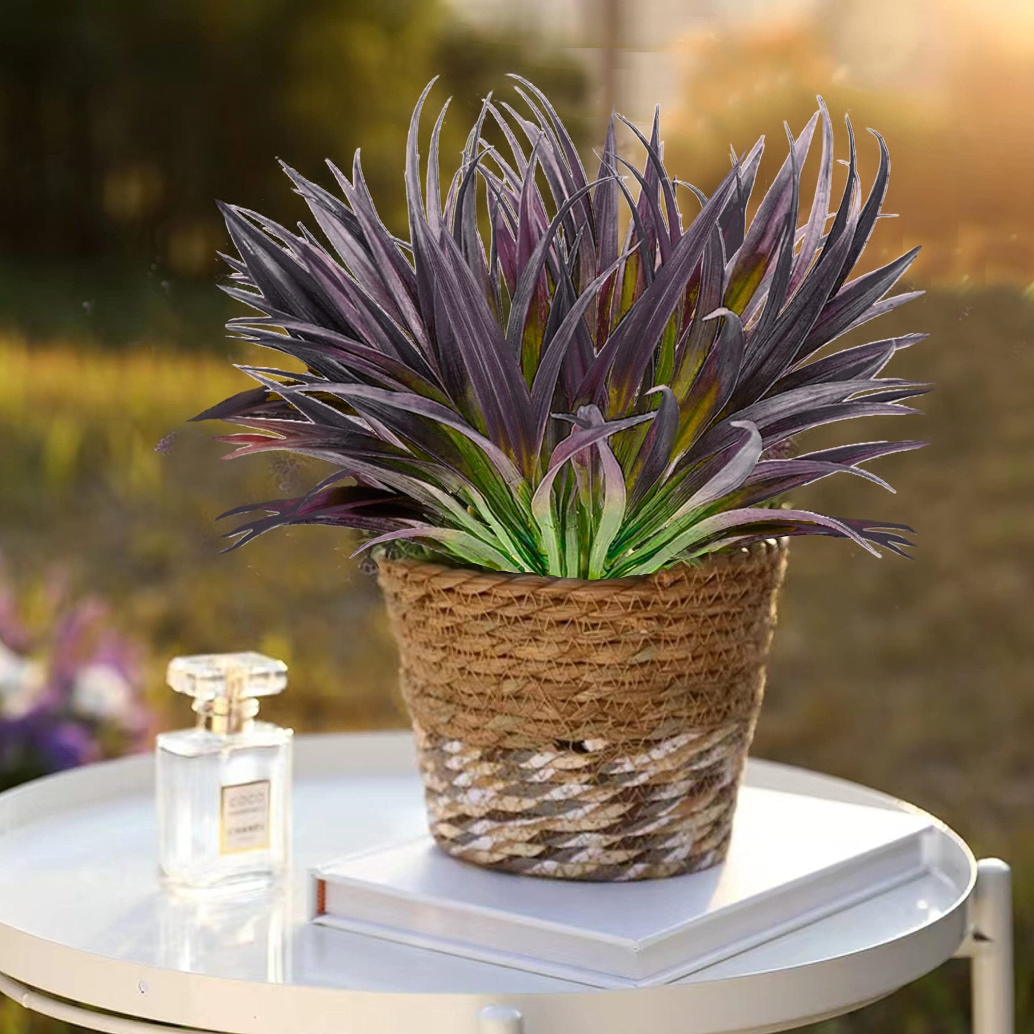 Artificial purple and green grass plant in a woven pot on a white table next to a perfume bottle.