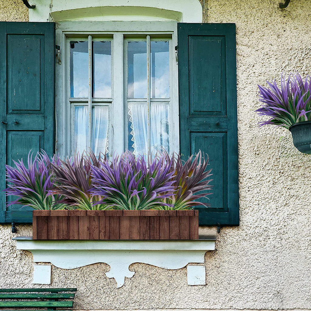 Artificial purple grass plants in a wooden planter box on a windowsill with dark green shutters.
