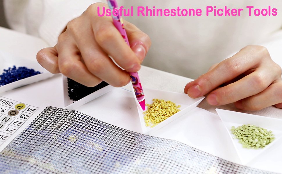 Close-up of hands using a pink rhinestone picker tool to pick up yellow beads from a white tray, with other trays of beads and a diamond painting grid visible.