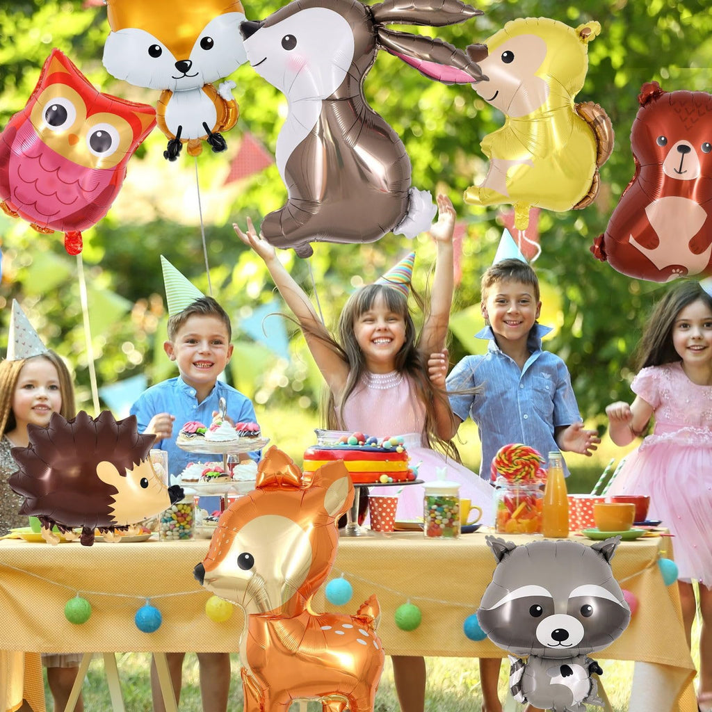 Children at a woodland-themed birthday party with assorted animal foil balloons floating above a table with cake and candy.