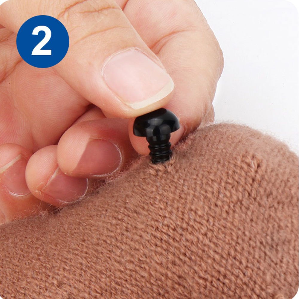 Close-up of a hand inserting a black plastic safety eye into brown knitted fabric, with a blue circle containing the number 2.