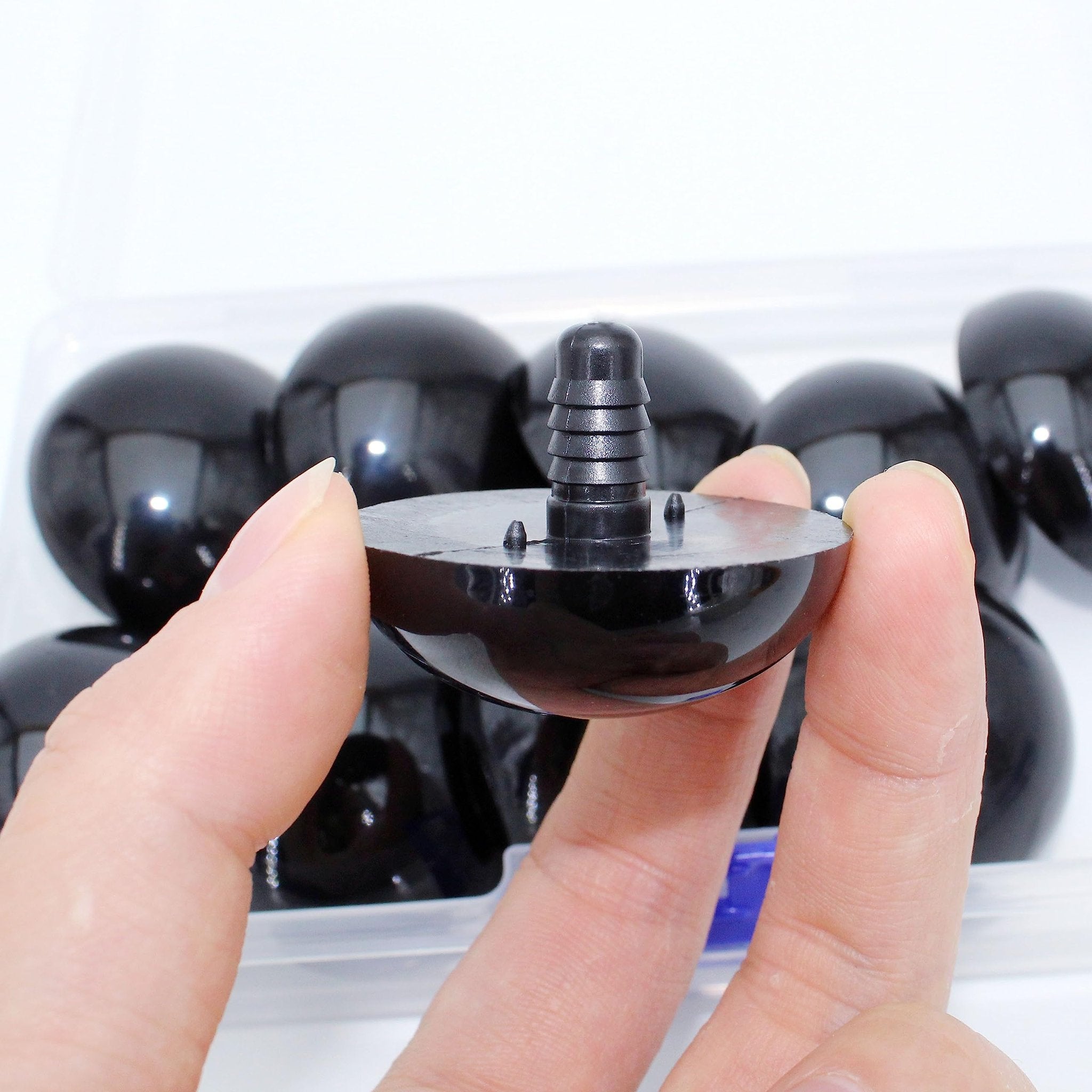 Close-up of a hand holding a black safety eye with a plastic washer stem, showing a row of similar eyes in a clear container.