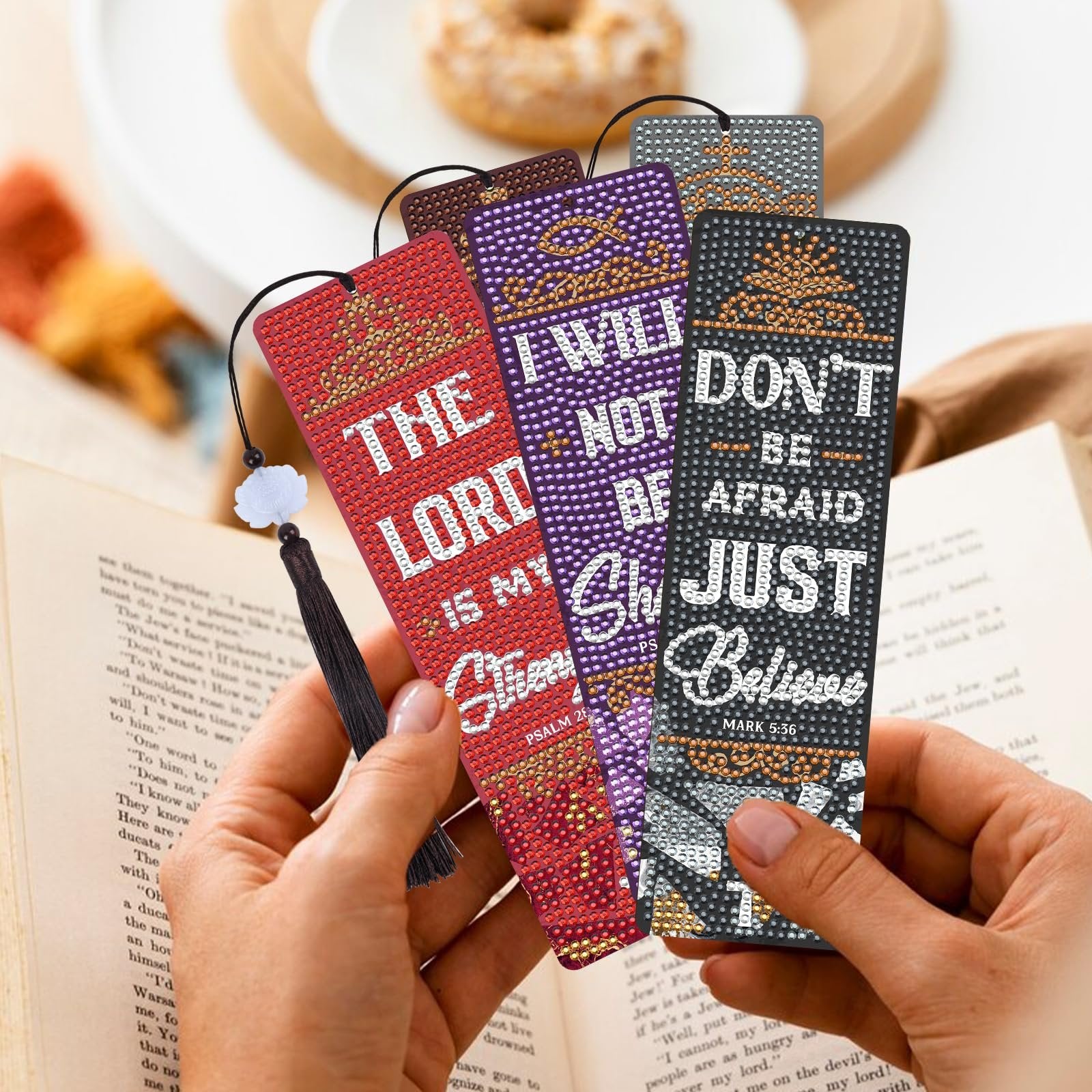 Hands holding four diamond art bookmarks with Bible verses and tassels, resting on an open book.