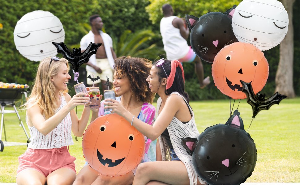 Three women toasting drinks at a backyard party with Halloween-themed balloons including a black cat, pumpkin, mummy, and bats.