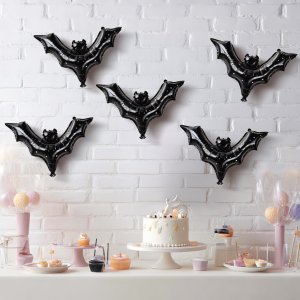 Five black bat foil balloons arranged on a white brick wall above a dessert table with a cake and cupcakes.