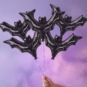 Hand holding a bunch of black bat-shaped foil balloons with glitter accents against a purple and pink gradient background.