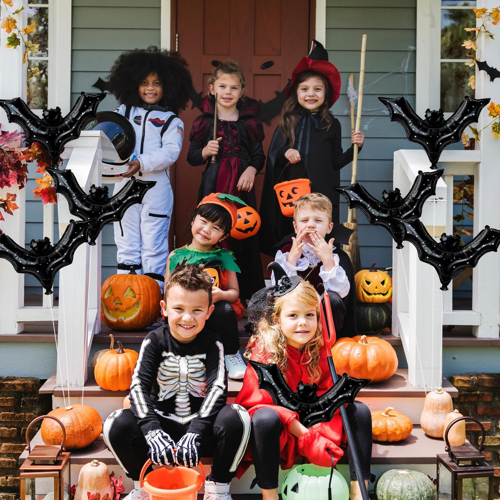 Children in Halloween costumes posing on porch steps with pumpkins and black bat balloons