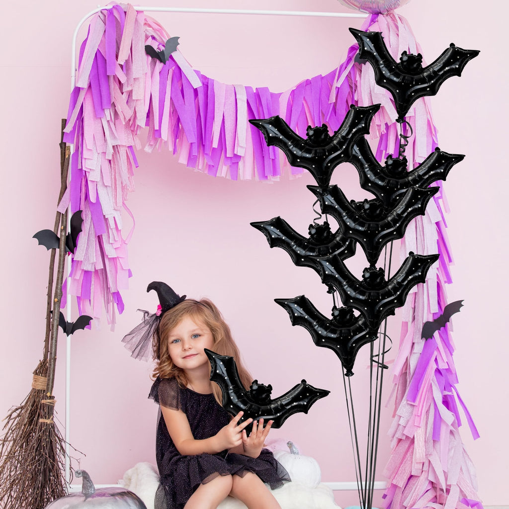 Young girl in witch costume holding black bat balloon with star pattern, surrounded by Halloween decor