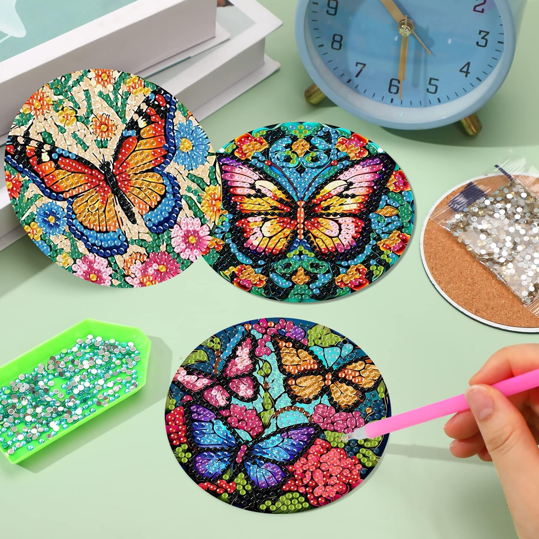 Diamond painting coasters with butterfly designs, tray of diamonds, and a pink applicator tool.