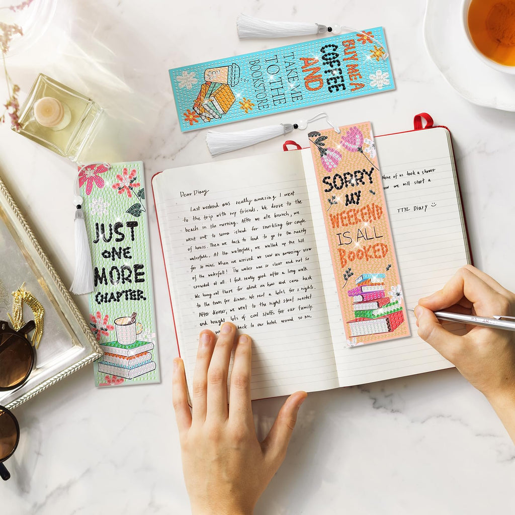 Diamond art bookmark kits with tassels on a notebook with handwritten entries and a cup of tea.