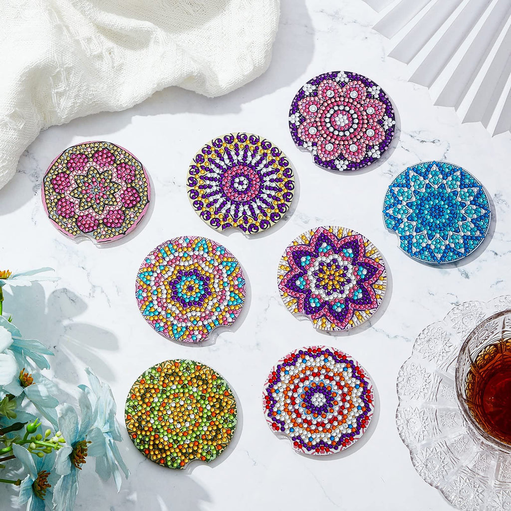 Eight diamond painting car coasters with mandala designs and cork pads on a marble surface with a white cloth and blue flowers.