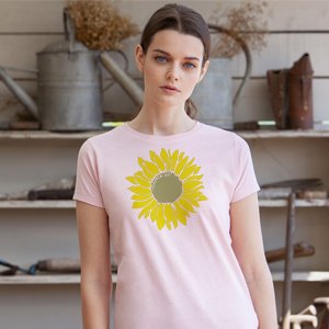 Woman wearing a pink t-shirt with a large yellow sunflower graphic on the front, standing in front of a rustic shelf with watering cans.