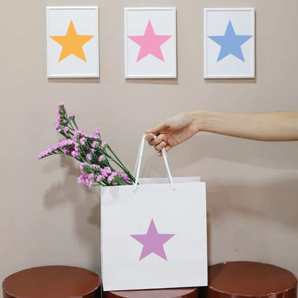 Hand holding a white gift bag with a purple star stencil on it, next to framed art with colorful stars.