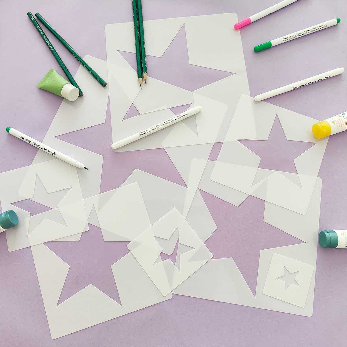 Various sizes of star stencils scattered on a purple surface with art supplies like pencils, pens, and glue.