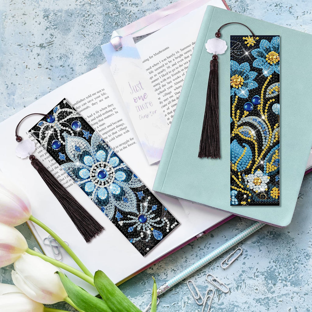 Two diamond art bookmarks with floral patterns and tassels are displayed on an open book and a notebook.
